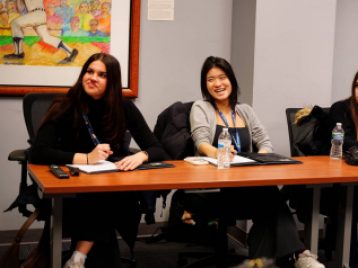 Three students sitting at a conference table, smiling and looking off camera, with part of a painting of a baseball player within the frame.